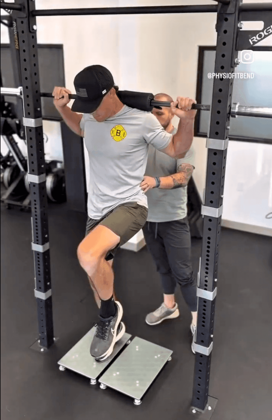 Front view of a client performing a barbell step-up on force plates as part of an ankle surgery rehabilitation program. He balances on the recovering ankle while driving the opposite knee up, with a trainer spotting from behind to ensure safety.