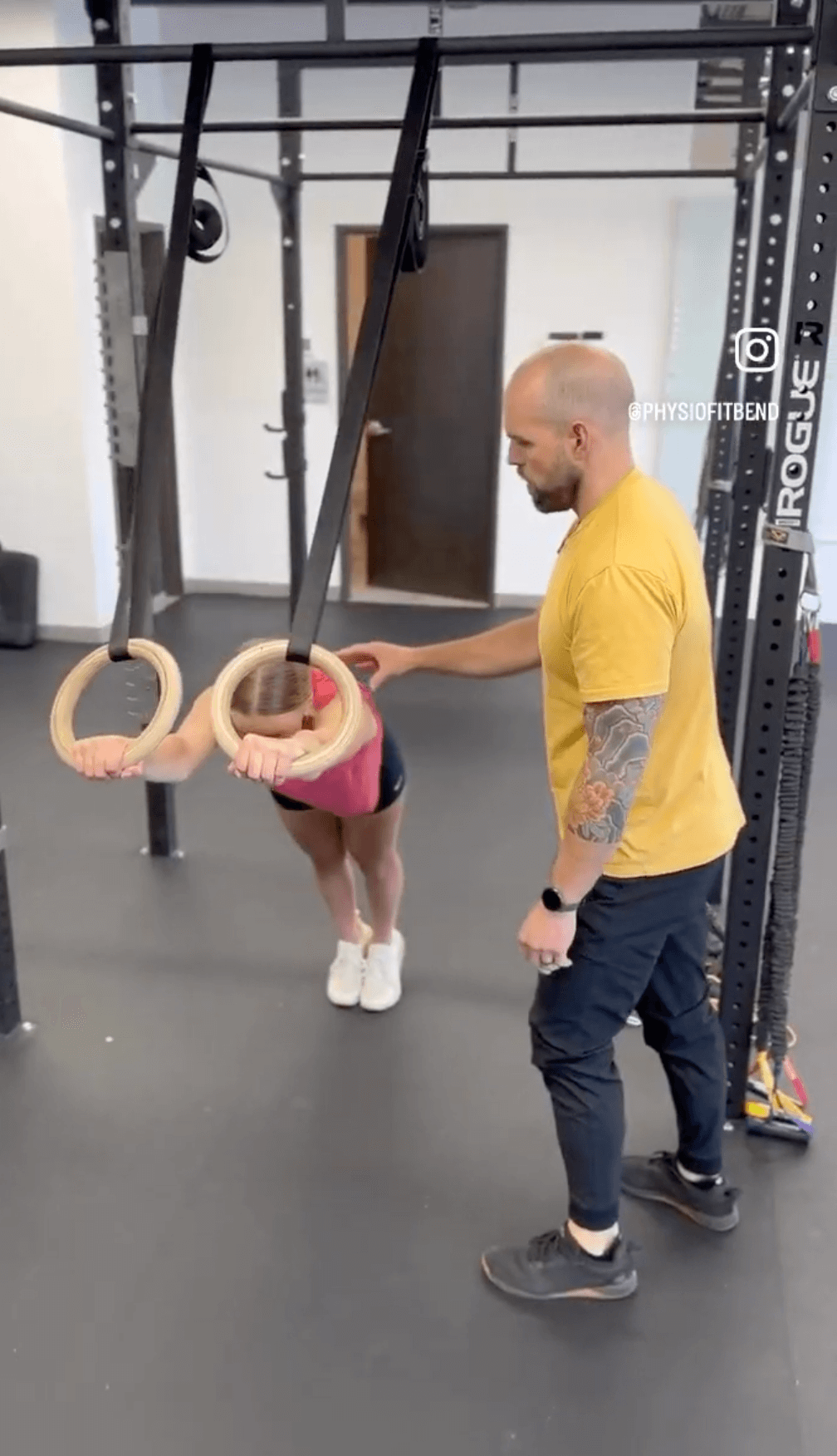 Woman performing an advanced gymnastic ring plank with arms extended, while a personal therapist provides tactile cues for proper back alignment.