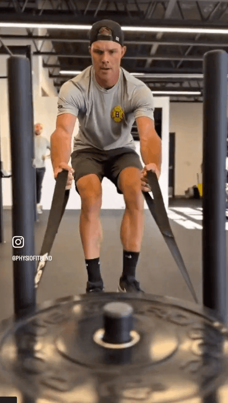 Head-on view of a man in a gym, leaning back and gripping two straps to pull a weighted sled across the gym floor.