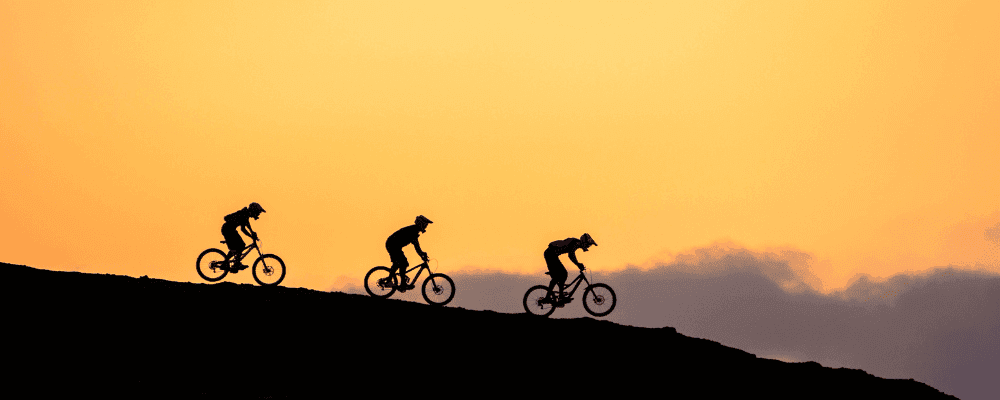 Silhouette of three men on bikes riding down a mountain against a sky at sunset.