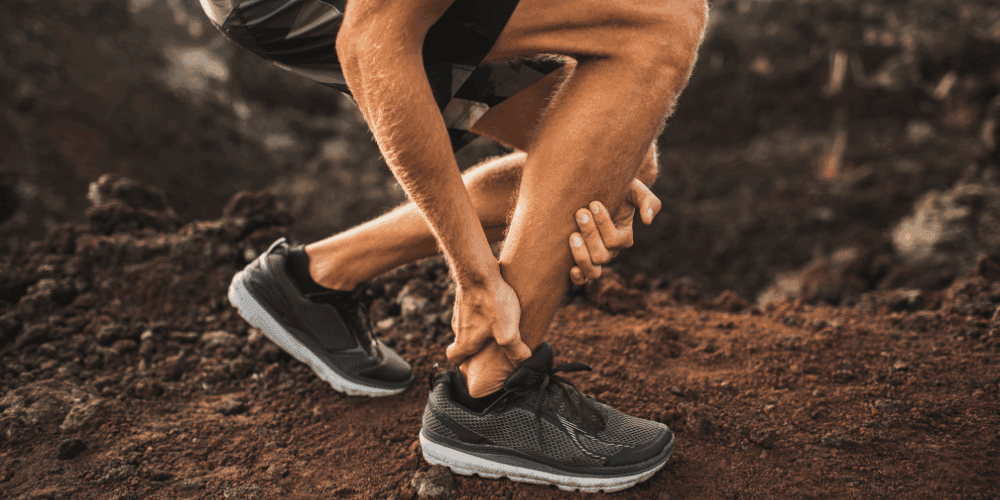 Man in running shoes on a trail outside bent down holding his ankle in pain.