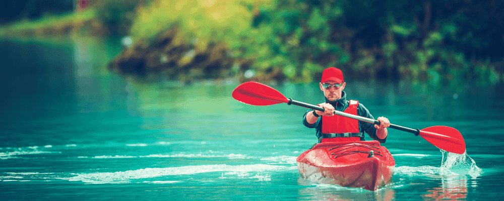Man in a red kayak with a red paddle and hat, kayaking down a river.