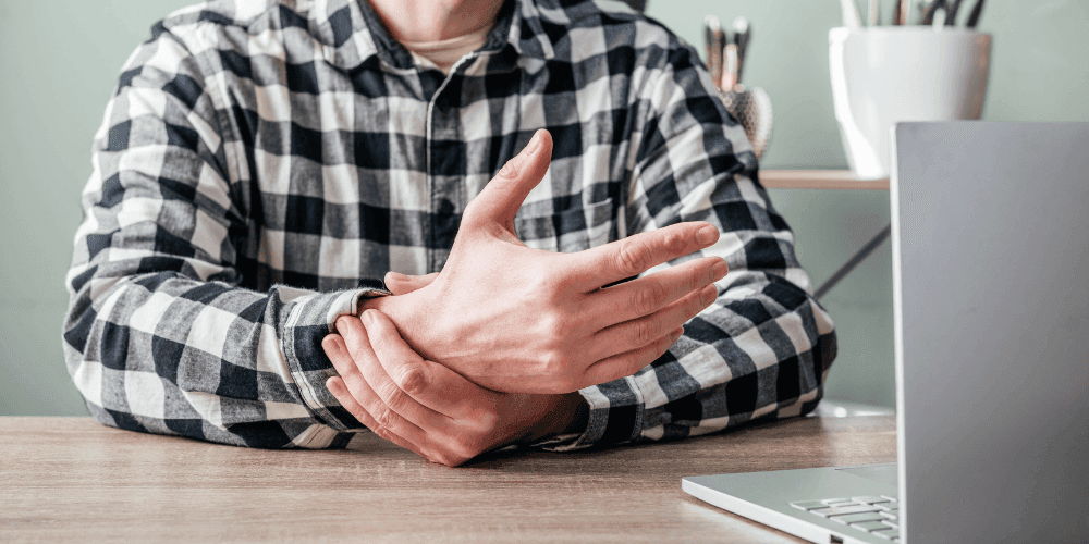 Man holding his wrist in pain at his desk in front of a laptop.