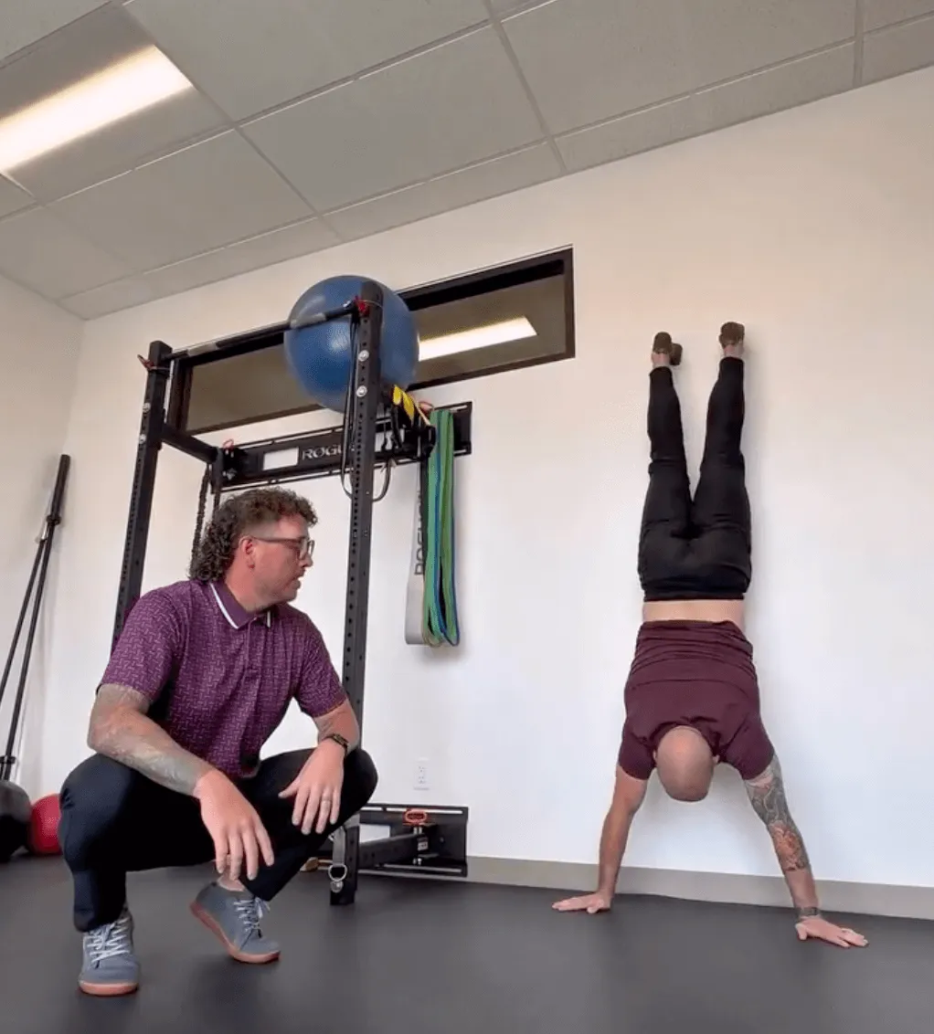 Rear view of a physical therapist performing a strict wall-facing handstand in the gym. A second therapist kneels nearby, observing spinal alignment and shoulder stacking.