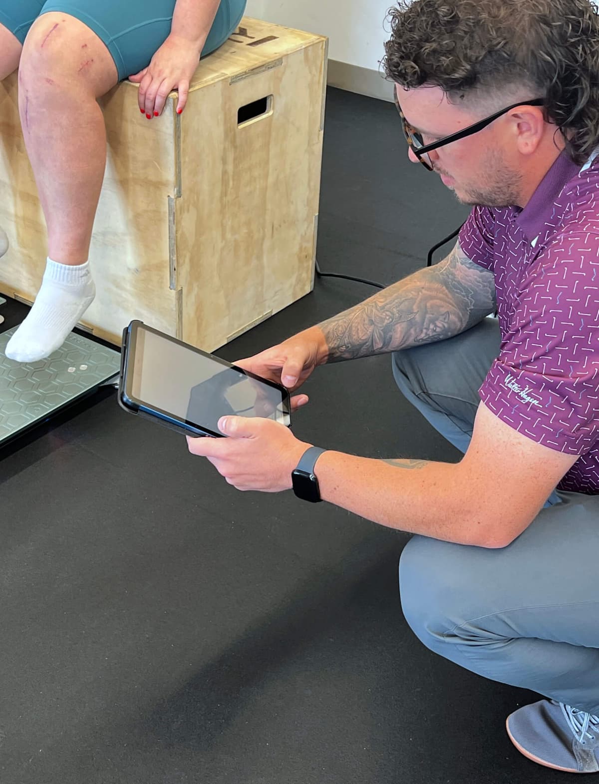 Woman sitting on a wooden box in a gym, showing a healing incision from ACL reconstruction surgery. A physical therapist squats next to her, reviewing strength metrics on a tablet.