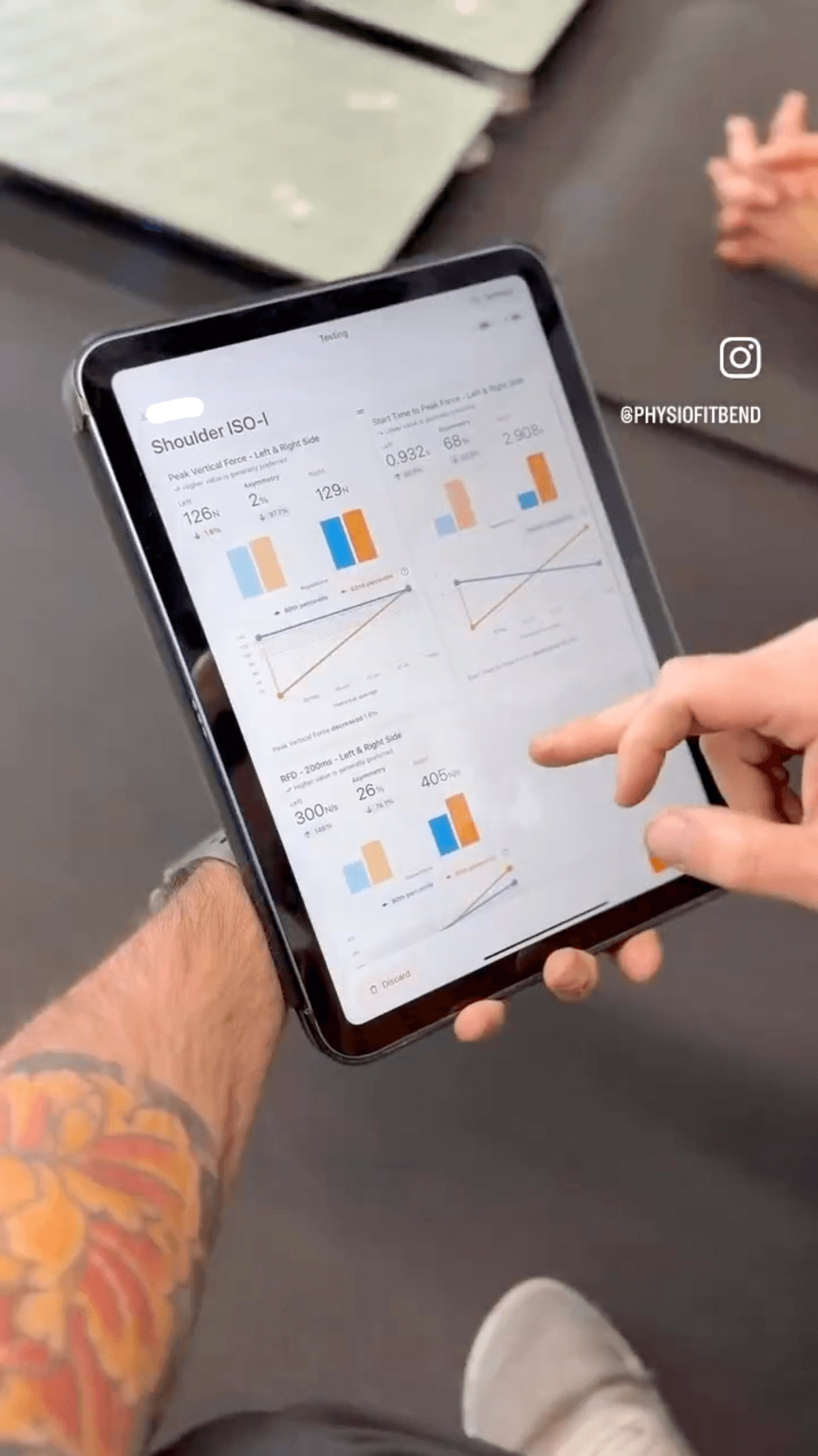 Close-up view of a trainer's tablet displaying Shoulder ISO-I strength graphs. The trainer's hand is pointing to a specific peak force metric on the screen, explaining the results to the client.